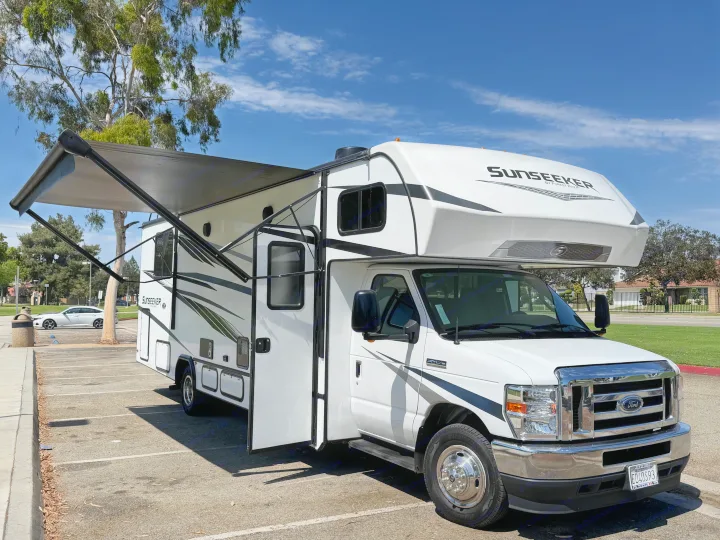 A white Sunseeker RV rental with its awning extended is parked in a sunny lot under a blue sky.. Forest River Sunseeker LE 2024
