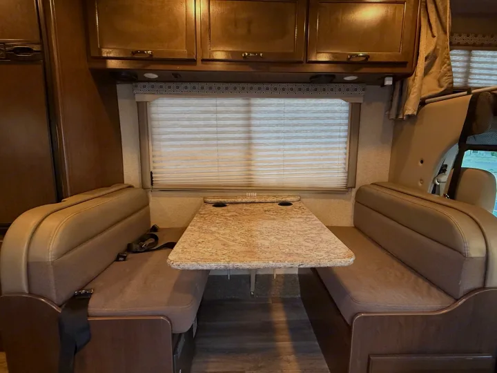 Inside an RV rental: a cozy dinette with tan benches, speckled table, wood cabinets, and blinds on the window.. Thor Four Winds 2018