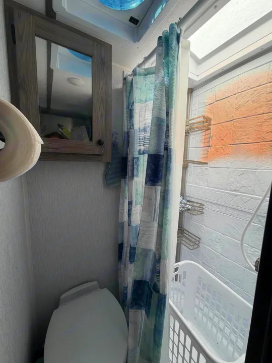 Compact RV rental bathroom featuring a toilet, shower with a patterned curtain, caddy, laundry basket, and mirrored cabinet.. Sunset Park RV Sun Lite 2025