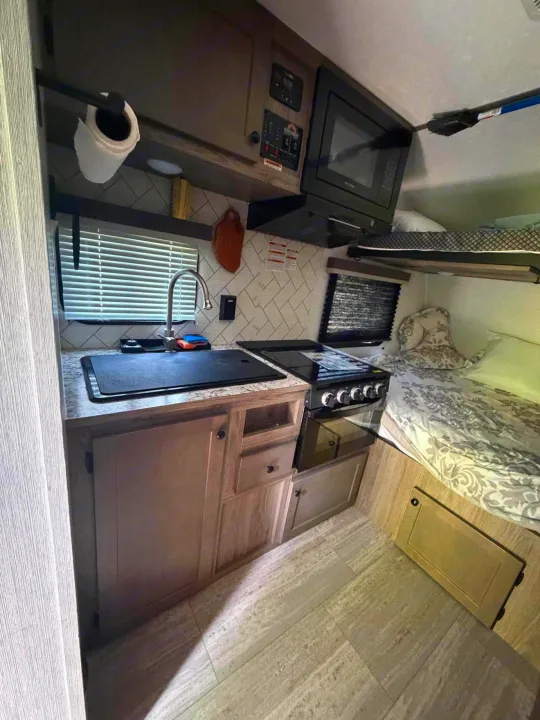 This RV rental interior shows a compact kitchen with sink, stove, microwave, plus a lower bed and an upper bunk.. Sunset Park RV Sun Lite 2025