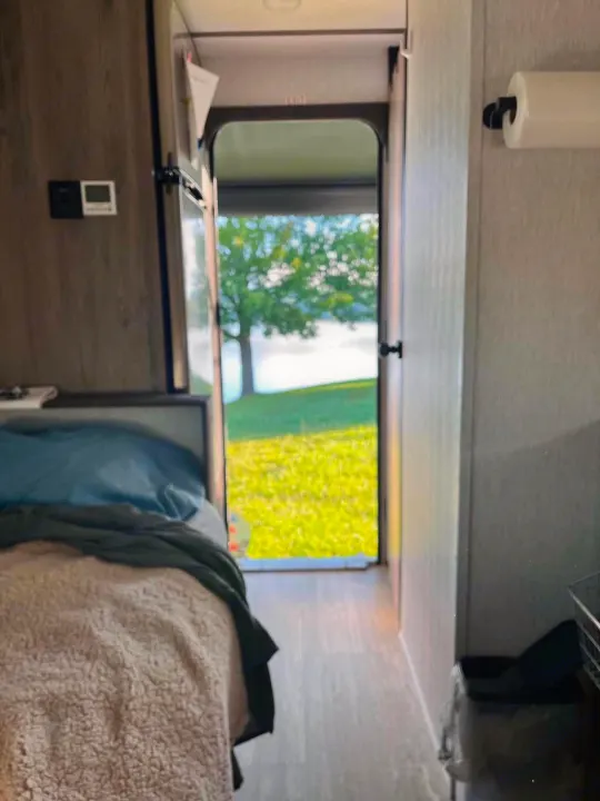 RV rental interior with a cozy bed and open door revealing a scenic lake view, perfect for camping.. Sunset Park RV Sun Lite 2025