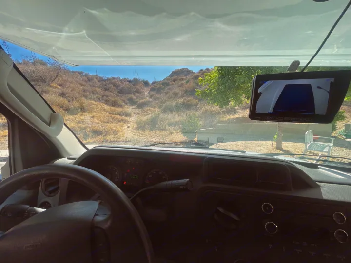 RV rental dashboard view reveals a dry, hilly landscape through the windshield, with a rearview monitor showing a campsite and chair.. Thor Majestic 2017