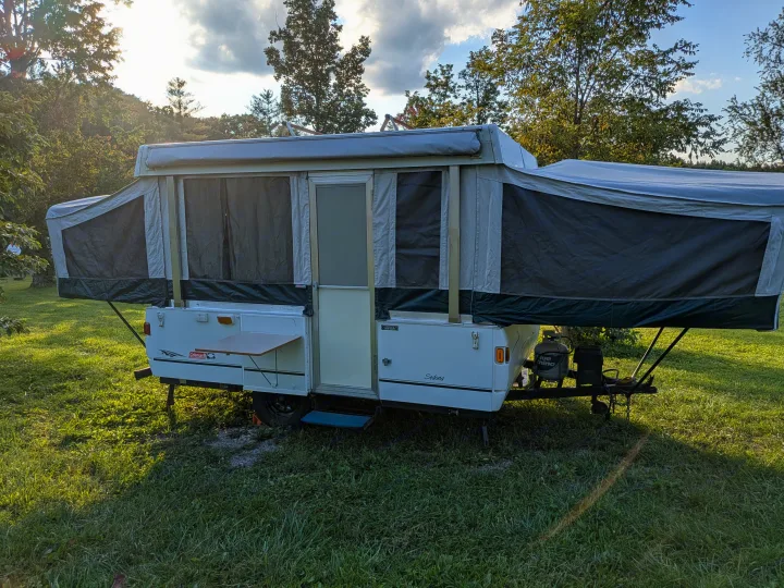 A white and gray pop-up camper (RV rental) with extended tents and table, set up on grass under evening sun.. Coleman Destiny Sedona 2002