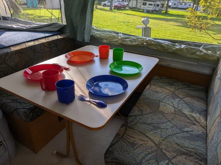 RV rental interior: A pop-up camper dining table with colorful dishware set for a meal, overlooking a sunny campground.. Coleman Destiny Sedona 2002