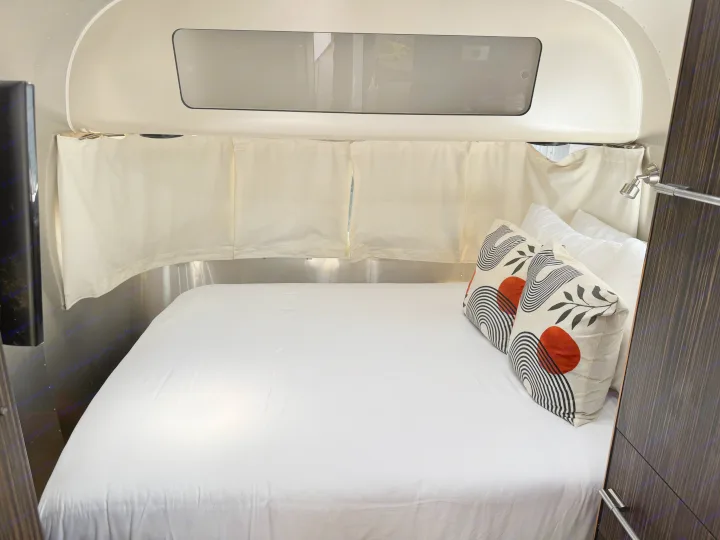Cozy RV rental bed with white sheets, modern red/black pillows, and window curtains, showcasing a compact and stylish interior.. Airstream International 2017