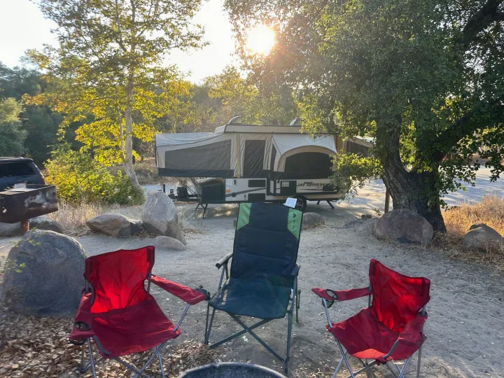 Pop-up RV rental camper at a sunny campsite, chairs arranged by the fire pit under sun-drenched trees.. Jayco 1209SC 2016