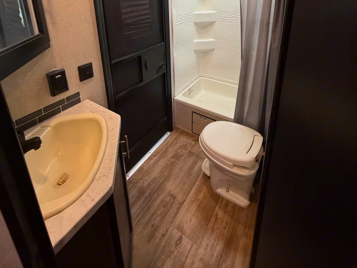 Inside an RV rental bathroom featuring a sink, toilet, and shower with a curtain, alongside wood-look flooring.. Forest River Cherokee Grey Wolf 2021