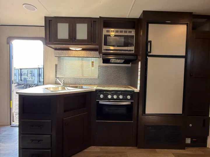 Modern RV rental kitchen with dark wood cabinetry, sink, stove, microwave, and Dometic fridge for your travels.. Heartland RVs Pioneer 2021