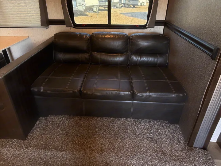 Inside an RV rental, a dark brown, three-seat faux leather couch with white stitching sits against a light wall, next to a window.. Heartland RVs Pioneer 2021