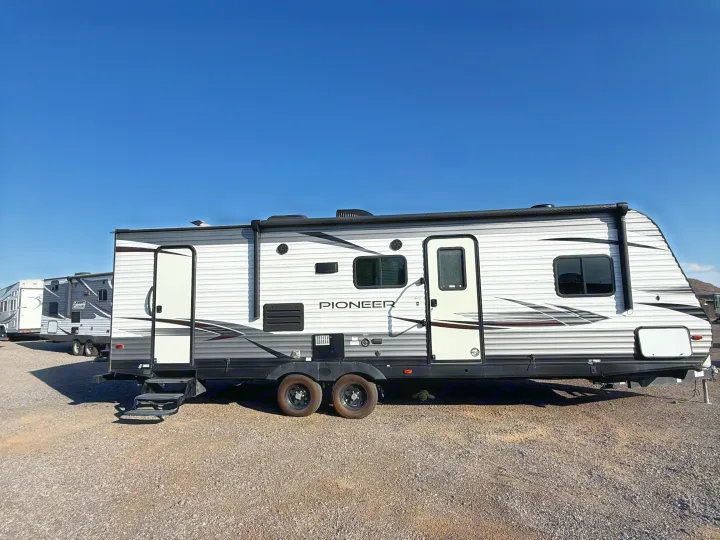 A Pioneer travel trailer, perfect for rv rental, parked on gravel under a clear blue sky. Steps are down; a small green toy is visible underneath.. Heartland RVs Pioneer 2021