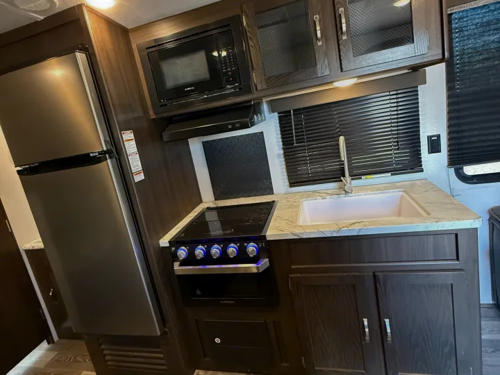 This RV rental kitchen features a refrigerator, microwave, stove, sink, and ample dark wood cabinets with marble-look counters.. Keystone RV Springdale 2021