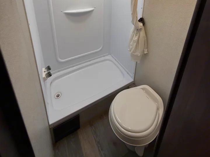 This RV rental bathroom features a compact white shower stall with a shelf, a toilet, and a tied-back shower curtain.. Keystone RV Springdale 2021
