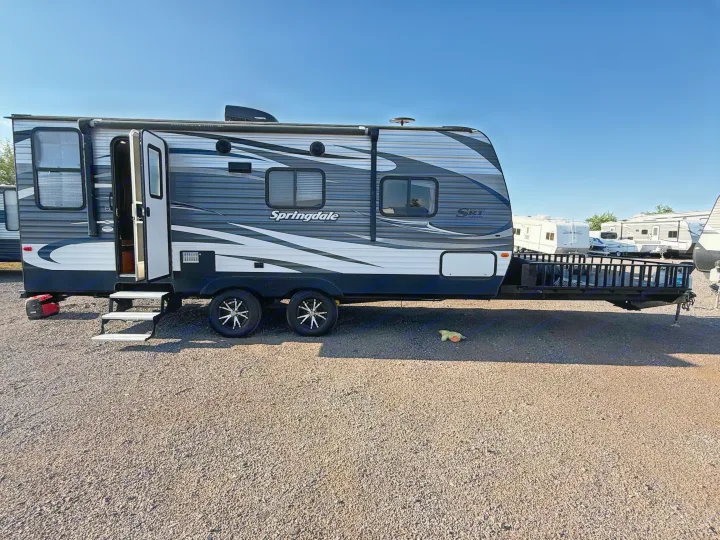 Springdale travel trailer RV rental, grey & white, door open with steps down, on a gravel lot under a clear blue sky.. Keystone RV Springdale SRT 2016