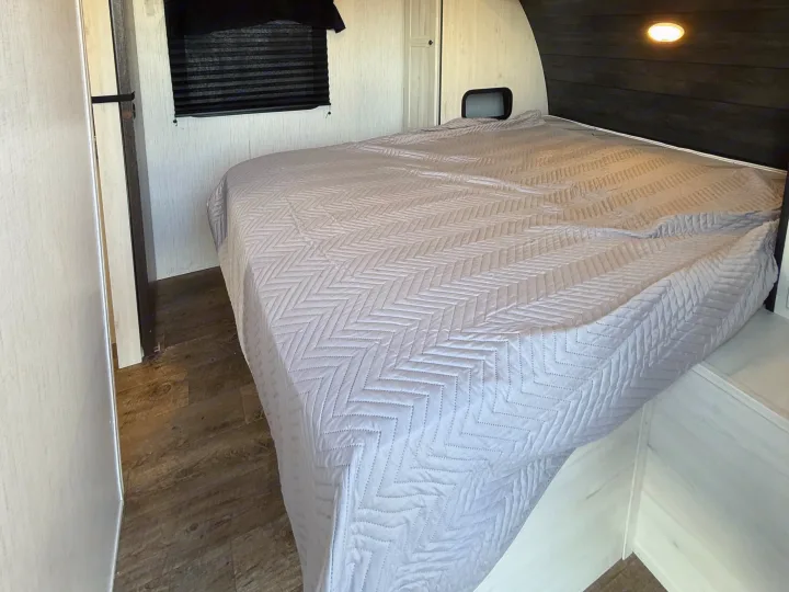 RV rental bedroom with a large, light grey quilted bed, wood paneling, and a window, offering a comfortable and neat interior.. Heartland RVs Trail Runner 2022
