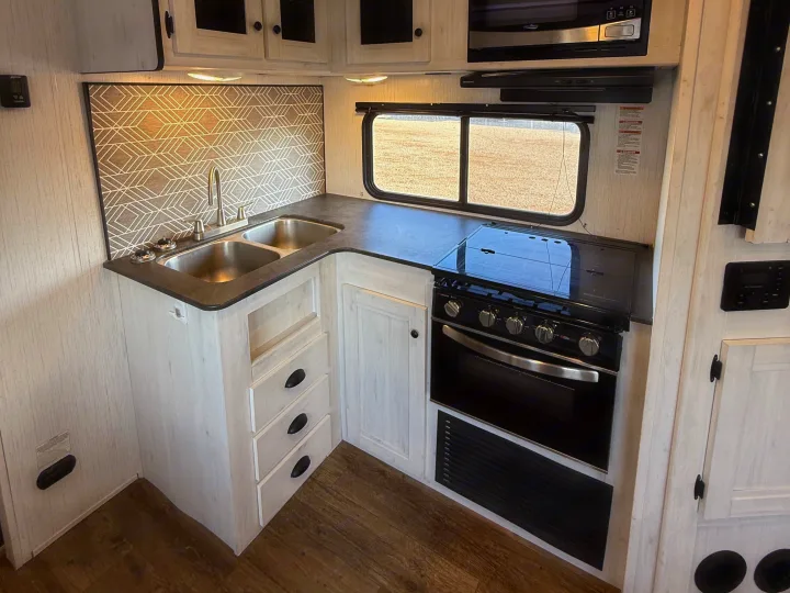 Inside a modern RV rental, this bright kitchen boasts a double sink, gas oven, and microwave, ready for meals.. Heartland RVs Trail Runner 2022