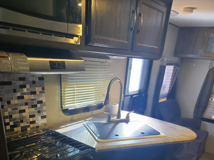 RV rental kitchen: A compact space with a stainless steel microwave, stove, tiled backsplash, sink, and overhead cabinets. Sunlight illuminates the counter.. Coachmen Apex 2017
