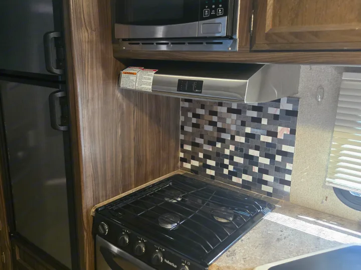 Compact RV rental kitchen with a stove, microwave, and refrigerator, featuring a stylish tiled backsplash and wood-grain walls.. Coachmen Apex 2017