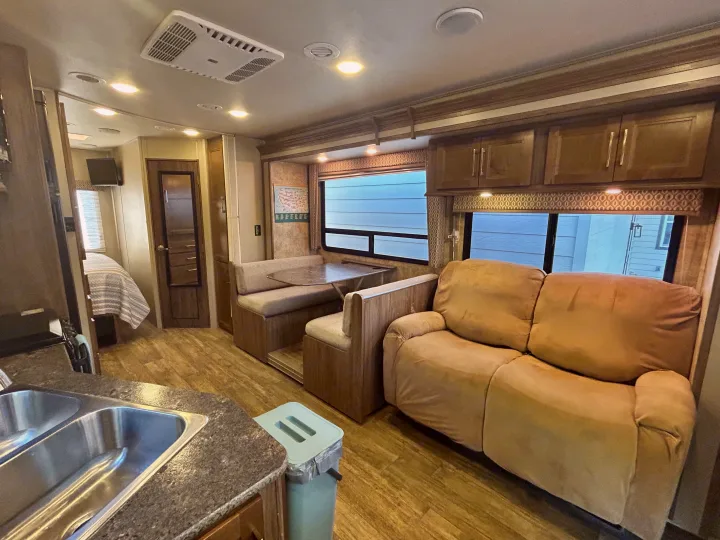 Inside this RV rental: a bright, wood-floored living area with tan sofa, dinette, and kitchen. Bedroom visible beyond.. Jayco Envoy 100 2018