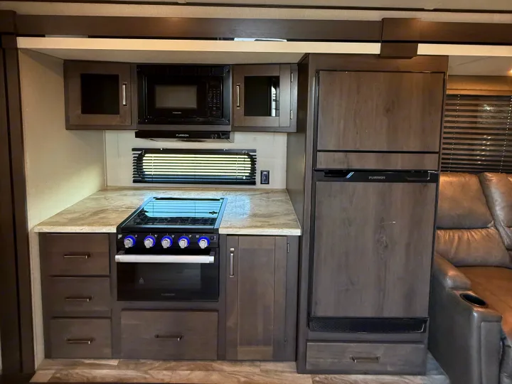 Compact RV rental kitchen featuring dark wood cabinets, light counter, Furrion microwave, stove, oven, and refrigerator.. Grand Design Transcend Xplor 2022