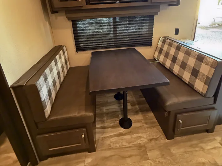 Cozy RV rental dinette featuring plaid benches, a dark table, under-seat storage, and a window with blinds.. Grand Design Transcend Xplor 2022
