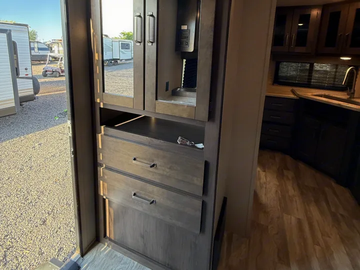 Modern RV rental interior showing dark wood cabinetry, drawers, a kitchen area, and an exterior view in the mirror.. Grand Design Transcend Xplor 2022