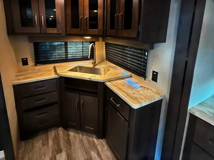 Modern RV rental kitchen interior with dark cabinets, marbled countertops, a stainless steel sink, and window blinds.. Grand Design Transcend Xplor 2022