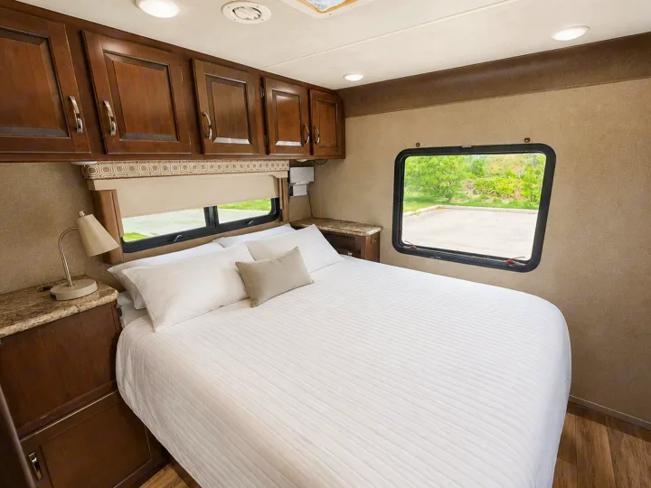 Cozy RV rental bedroom featuring a comfortable queen bed with white bedding, wooden overhead cabinets, and windows with outdoor views.. Thor Four Winds 2020