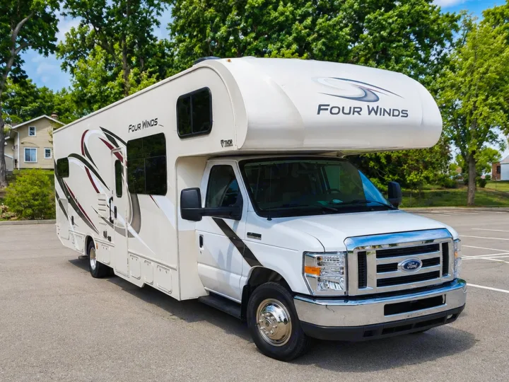 White Four Winds RV rental on a Ford chassis, parked in a paved lot with green trees and a house in the background.. Thor Four Winds 2020