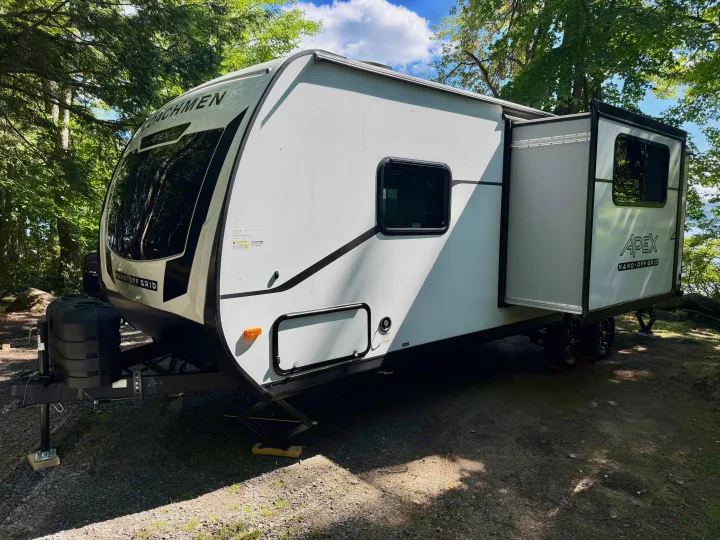 Shaded RV rental, a white Coachmen travel trailer with slide-out, parked at a peaceful, wooded campsite.. Coachmen Apex Nano 2026