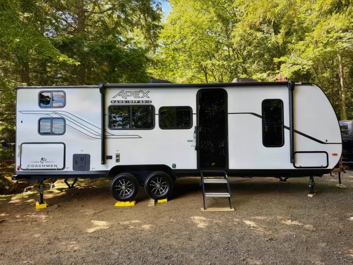 This white Coachmen Apex Nano RV rental is set up at a shaded campsite, surrounded by tall green trees, perfect for camping.. Coachmen Apex Nano 2026