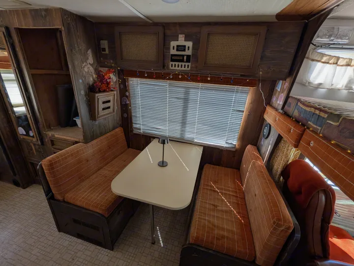 A classic RV rental interior featuring a cozy orange dinette, rustic wood paneling, blinds on the window, and an upper bunk bed.. Chevrolet Vanguard 1980