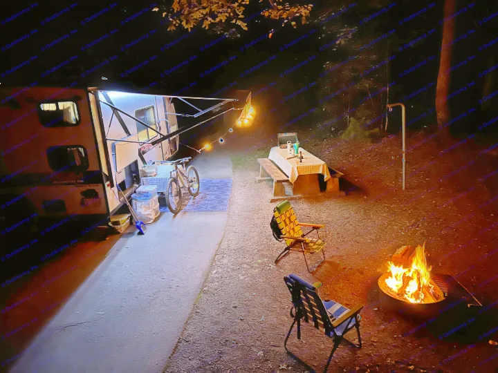 RV rental campsite at night with a cozy campfire, illuminated awning, picnic area, bicycle, and camping chairs.. Forest River Wildwood FSX Platinum 2021