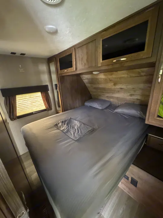 Inside an RV rental, this bedroom features a comfy grey bed, wood cabinets, and a window for natural light.. Dutchmen Coleman Light 2019