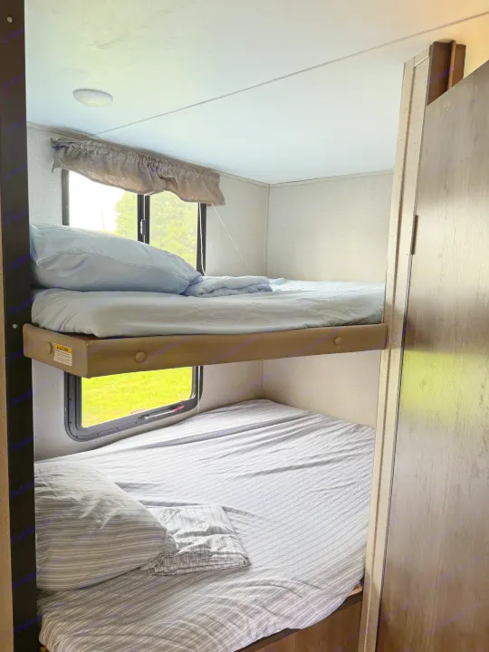 RV rental bunk beds: a cozy interior view with two stacked sleeping areas, light-colored sheets, and windows revealing greenery.. Dutchmen Coleman Light 2019