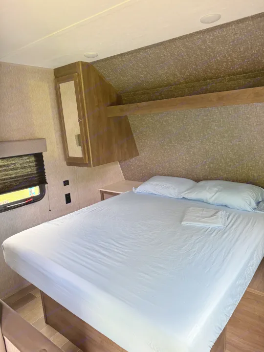 A compact rv rental bedroom showcasing a bed with light blue sheets, pillows, a window, and wood cabinetry.. Dutchmen Coleman Lantern LT 2020