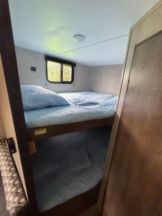 View inside an RV rental showing two bunk beds with light blue bedding, a window, and a charging port on the wall.. Dutchmen Coleman Lantern LT 2020