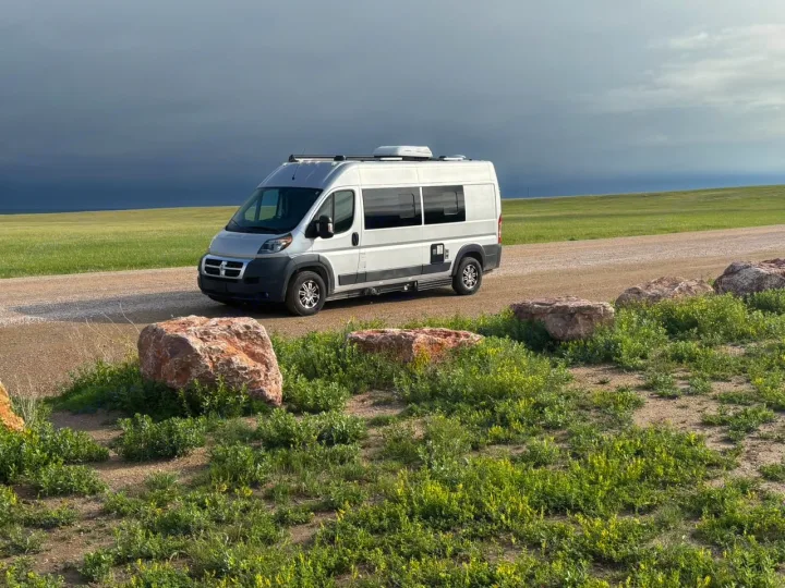Silver RV rental parked on a dirt road in a green field under a stormy sky, with rocks and lush foliage in the foreground.. Hymer Carado 2018