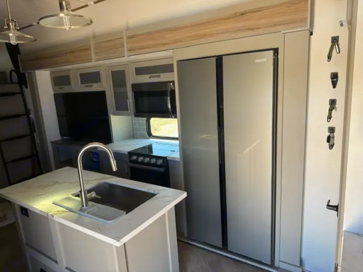 Modern RV rental kitchen featuring stainless steel appliances, island sink, TV, overhead cabinets, and a ladder to a loft.. Keystone RV Sprinter 2025