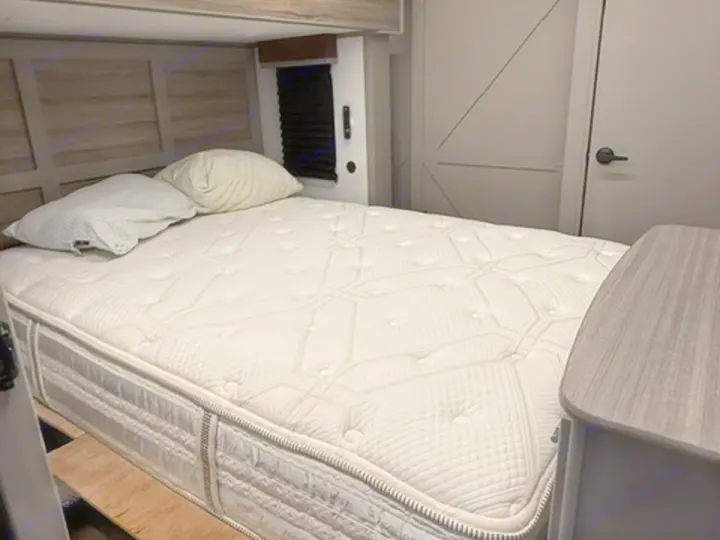 Comfortable RV rental bedroom with a white mattress, pillows, wooden headboard, and a small cabinet, offering a cozy sleep space.. Keystone RV Sprinter 2025