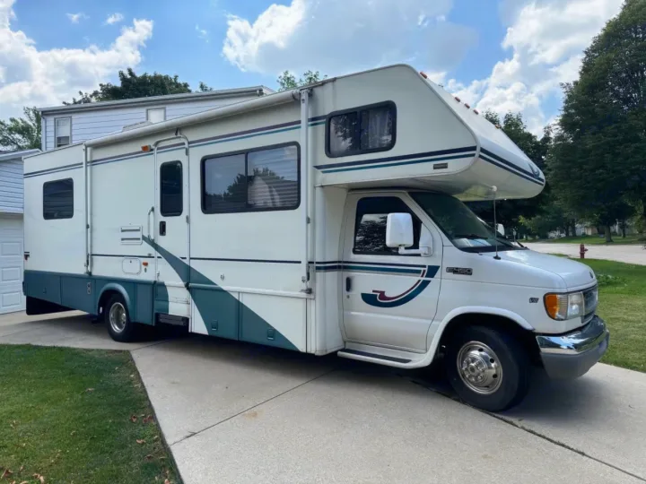 White Class C RV for rv rental, parked on a driveway. This motorhome features teal and maroon stripe accents.. Winnebago Itasca 2001