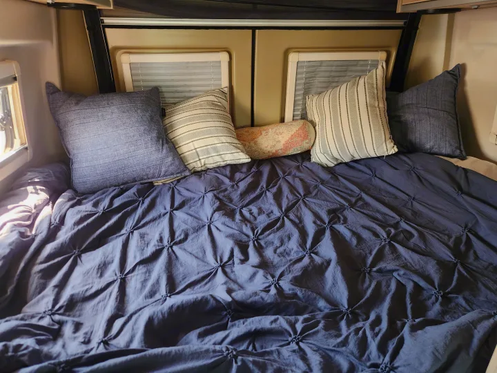 Comfortable sleeping setup in an RV rental, featuring a blue tufted comforter & assorted throw pillows.. RAM ProMaster 3500 2016