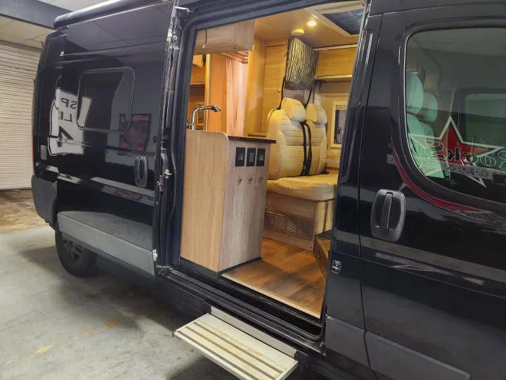 Open door view of a black RV rental interior: modern kitchen, comfortable seating, and wood finishes ready for your next trip.. RAM ProMaster 3500 2016