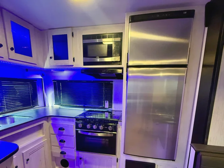 RV rental kitchen: blue accent lights illuminate stainless steel fridge, microwave, oven/stove, sink, and wood cabinets with blinds.. Heartland RVs Trail Runner 2022