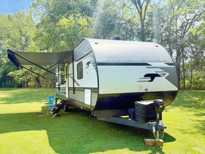 Trail Runner travel trailer, ideal for an RV rental. Parked on green grass, awning extended, with trees in a sunny outdoor setting.. Heartland RVs Trail Runner 2022