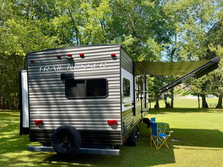 This Trail Runner travel trailer RV rental, with its awning and chairs, is parked in a sunny, tree-filled campground.. Heartland RVs Trail Runner 2022