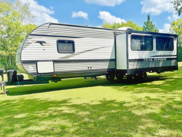 This Trail Runner travel trailer rv rental is parked on a sunny, grassy spot amidst trees, ready for your next outdoor adventure.. Heartland RVs Trail Runner 2022