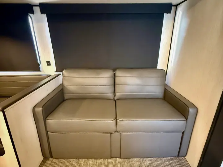 RV rental interior featuring a light brown two-seater sofa, dark window blinds, and a glimpse of a dinette on the left.. Heartland RVs Trail Runner 2022