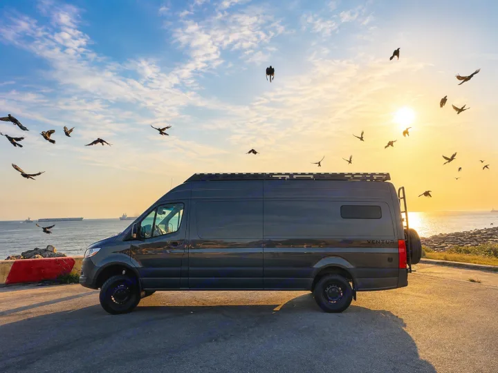 This dark gray Ventur camper van, perfect for RV rental trips, is parked by the ocean at sunset with many birds in the sky.. Mercedes Sprinter 2025