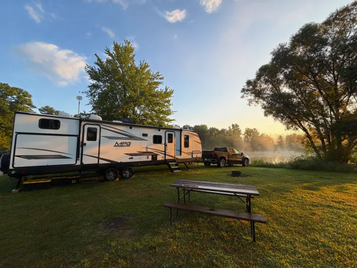 Sunrise at an RV rental campsite: travel trailer, truck, picnic table, fire pit, and a misty lake.. Cruiser RV MPG Ultra Lite 2022