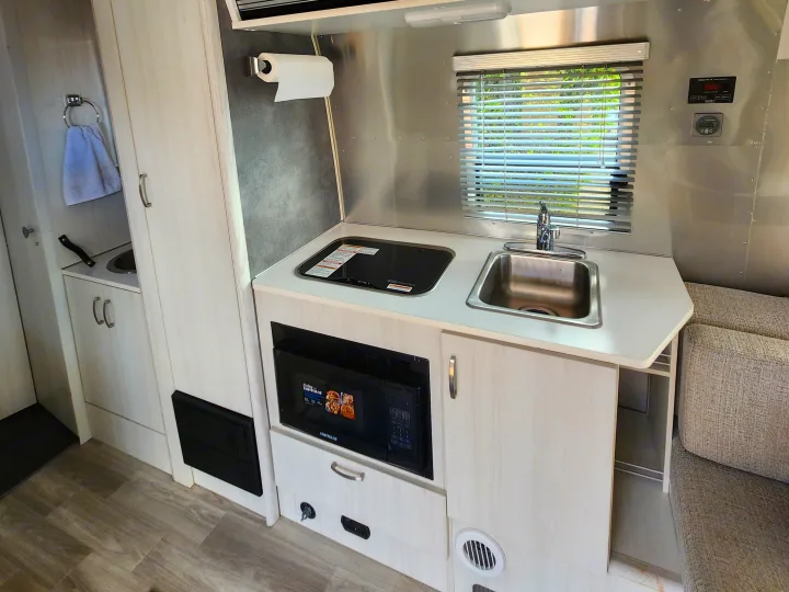 This RV rental's compact kitchen boasts a sink, stovetop, microwave, ample cabinets, and a window for scenic views.. Airstream Bambi 2023